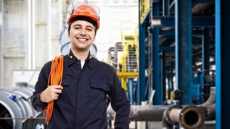 A smiling industrial worker in a hard hat, safety glasses, and navy coveralls holds an orange safety harness in a bright factory filled with pipes and machinery.