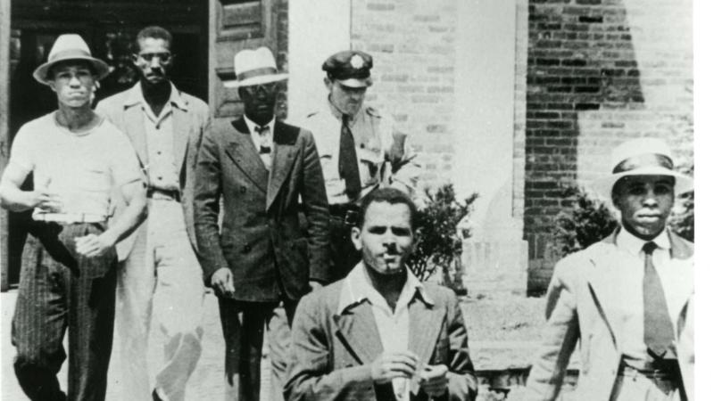 A black and white photo of the five African American men who participated in the library sit-in being escorted by a police officer from the library. Four of the five men are wearing a suit and tie.