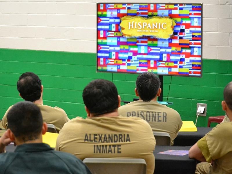 backs of five inmates, seated and wearing tan or green jumpsuits, looking at a large televsion screen with colorful flags displayed