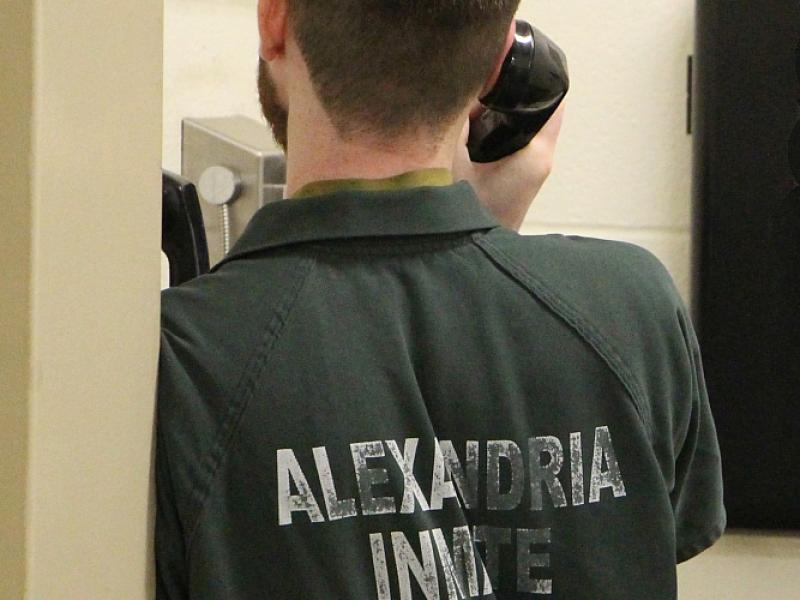 view from behind of an inmate wearing a green jumpsuit that says "Alexandria Inmate" and holding a phone to his ear