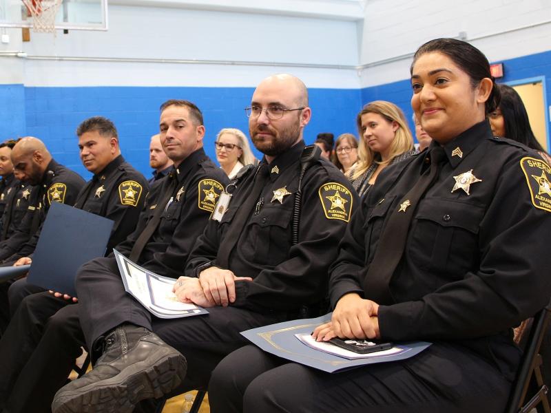 seven deputies in blue uniforms seated and looking forward with civilians visible in background