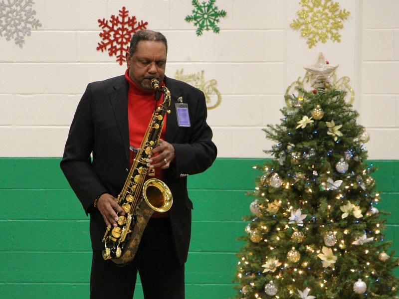 man wearing a black suite and red shit playing the saxophone with a Christmas tree and holiday decorations in the background