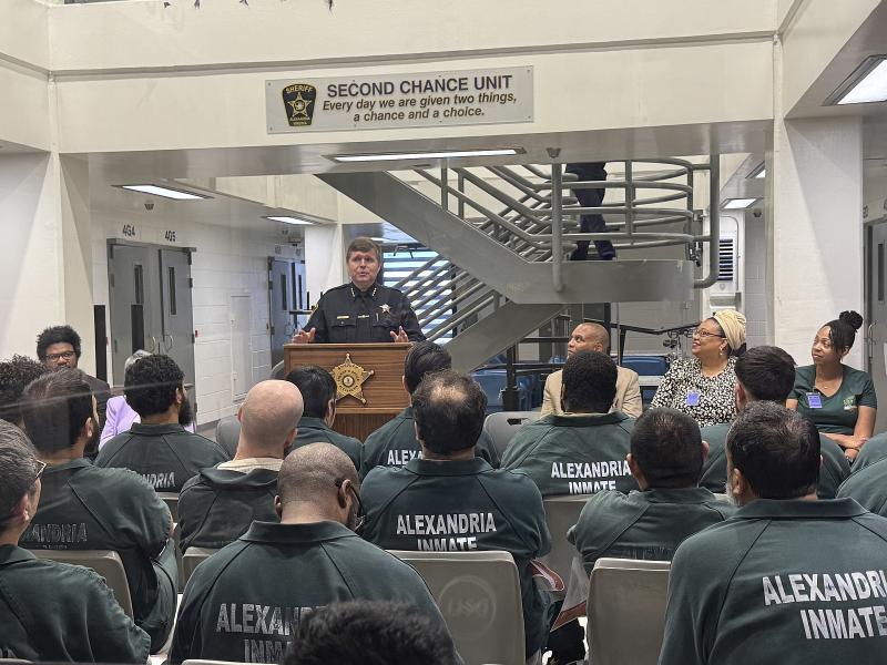 Sheriff standing at podium in front of several seated inmates wearing green jumpsuits and visible from the back