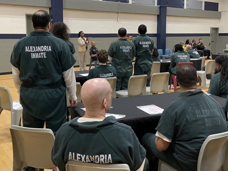 Several inmates in green jumpsuits seated in a gymnasium visible from behind with two guest speakers at a podium in the background