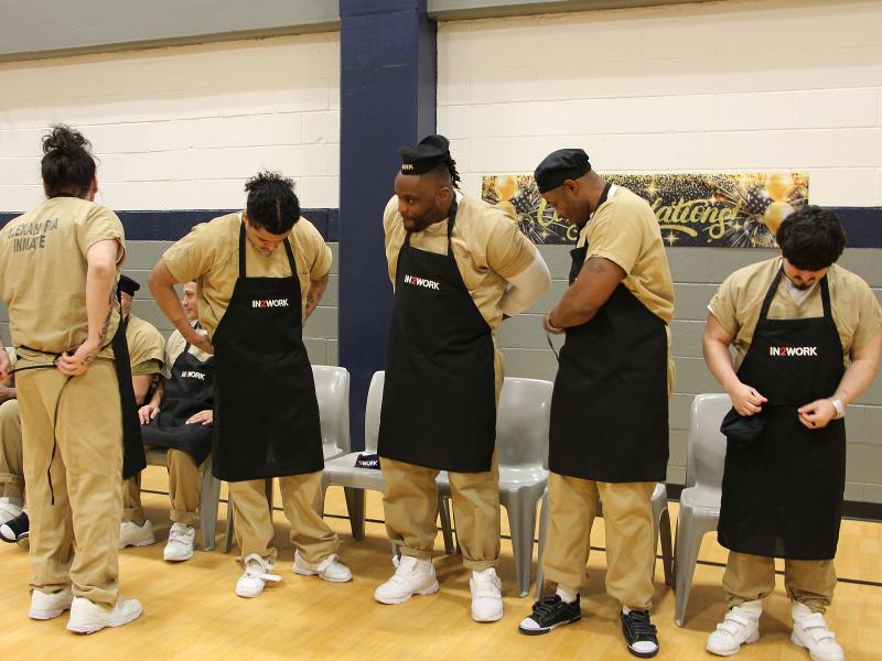 Five inmates in tan uniforms putting on black aprons