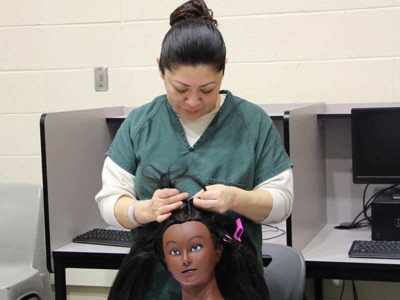 inmate in green uniform braiding the hair of a mannequin head