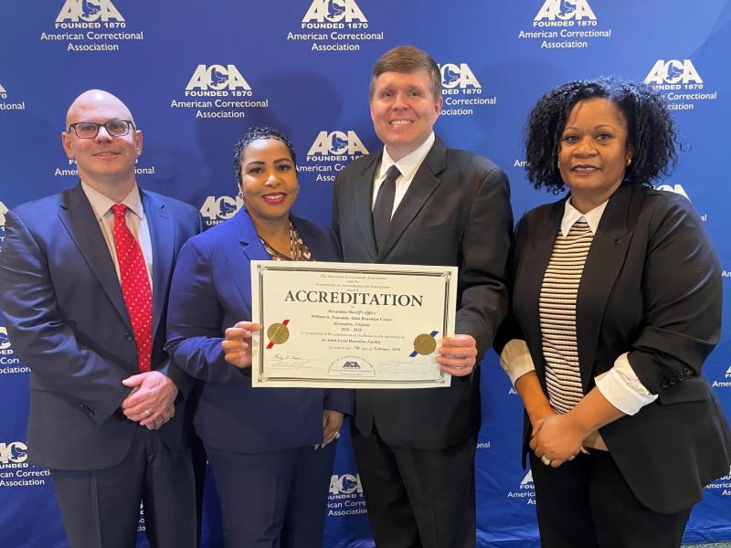 Four corrections executives in business attire standing next to each other with two in the center holding a certificate