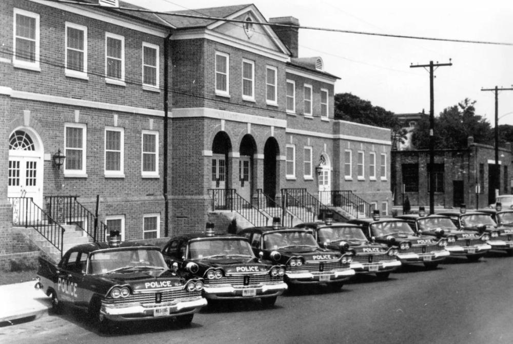 A black and white photo of a row of dark cars with POLICE written on the front of them in white letters, parked outside of a brick building.