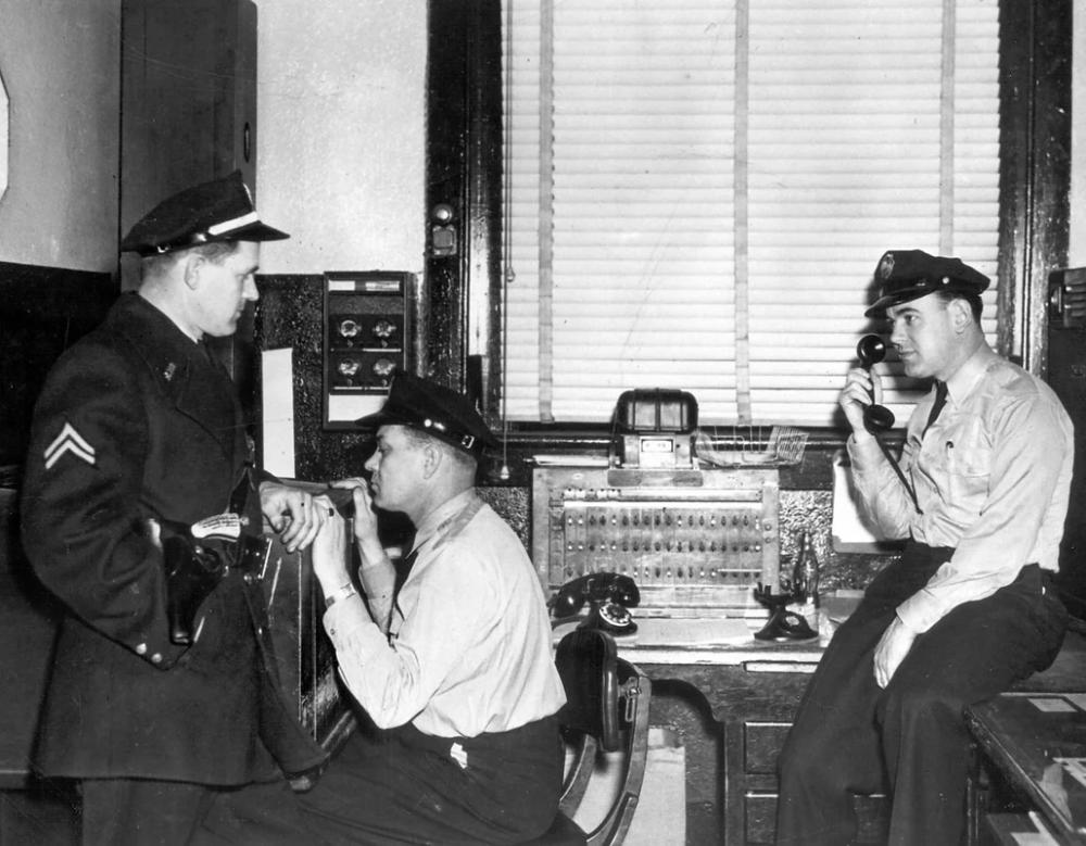 A black and white photo of three young white men in police uniforms. One is lounging against a desk, one is sitting, and the other is on the opposite side half-sitting on the desk and holding a phone receiver near his ear.