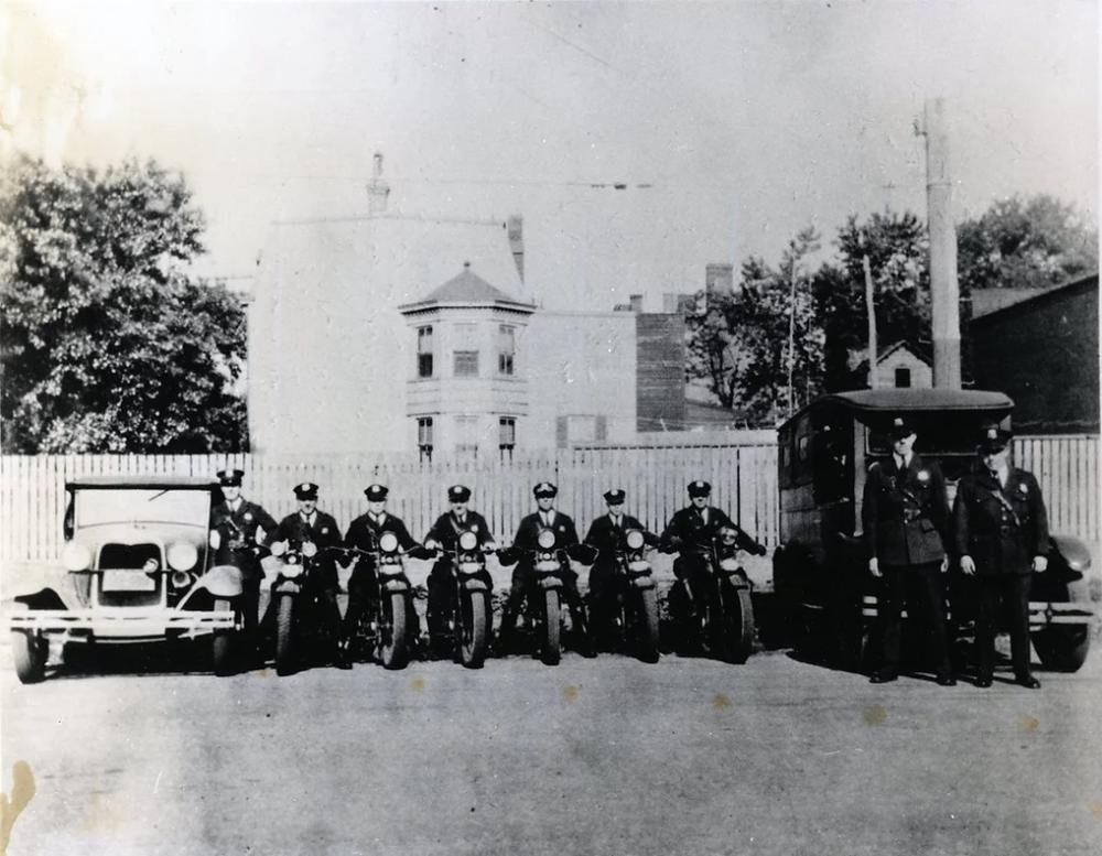 A black and white photo of seven men in police uniforms sitting on motorcycles, poised as though to drive toward the camera. On either side of the group of motorcycles is an old-fashioned car. In front of the car on the left are two more uniformed men, also facing the camera. In the background is a picket fence and an old-fashioned building.