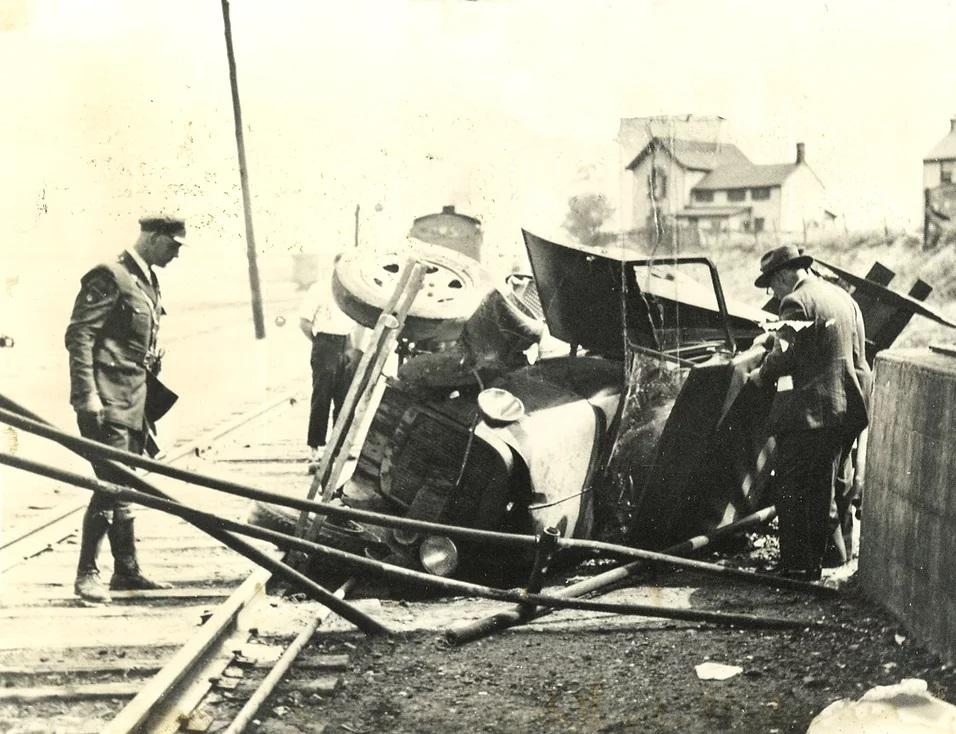 Two men, one in a suit and one in a uniform, inspect the wreckage of an old-fashioned car. The car is on its side next to a railroad track, badly damaged, and the windshield is broken.