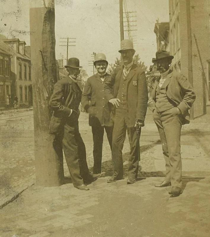 A sepia-tone photograph of four white man standing casually on a sidewalk. On the left, the man is leaning back against a telephone pole. To his right is a younger man standing slightly behind the groups and wearing a rounded policeman's hat/helmet. To the right of him is a tall man also wearing the rounded hat/helmet, and with a police badge on his jacket and a weapon in his hand. The fourth man is at the far right with his hands in his pockets and has a large moustache.