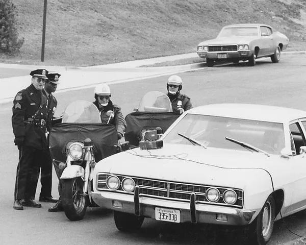 A black and white photo of two men in police uniforms standing beside two officers seated on motorcycles beside a light sedan. All are looking at a piece of equipment on the hood of the sedan. The photo appears to have been taken in the 1970s.