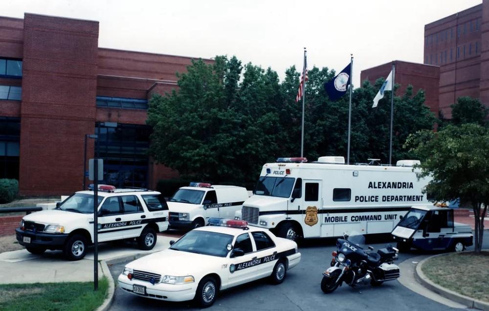 A color photo of a parking lot with a collection of APD vehicles (from the top, clockwise): mobile command unit, small utility vehicle, sedan, SUV, box van in front of a dark red brick building