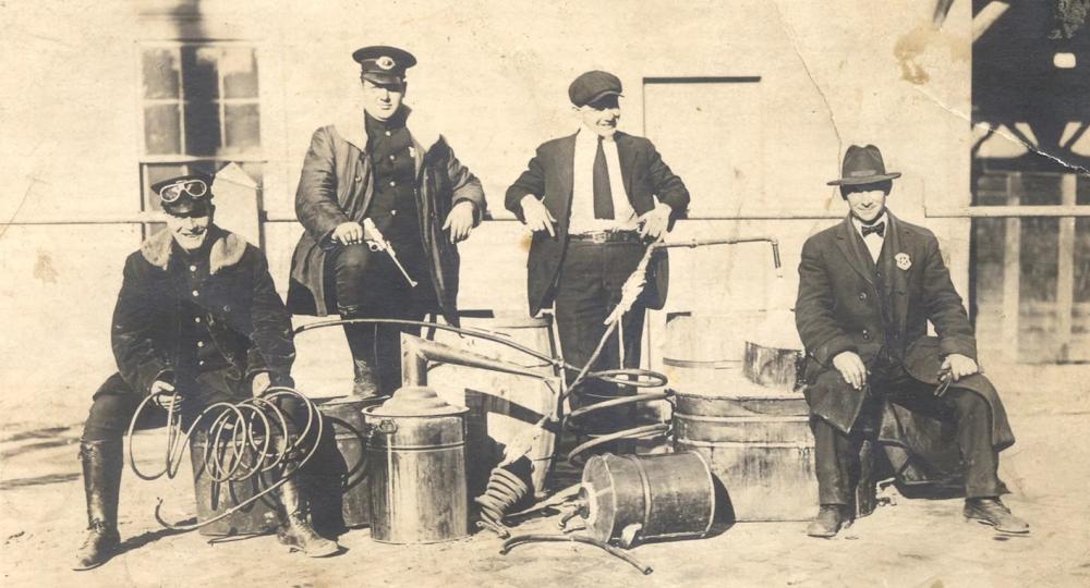 A sepia-toned photograph of four men with a pile of metal canisters and tubs, with two men standing behind the equipment and two sitting on the pile. The photo appears to have been taken during the Prohibition.