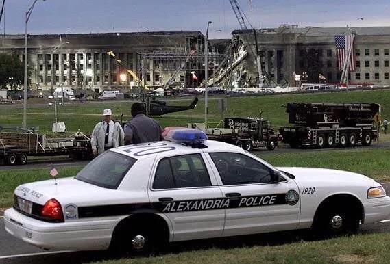 An Alexandria Police car is parked with two men standing near it and the damaged Pentagon in the background