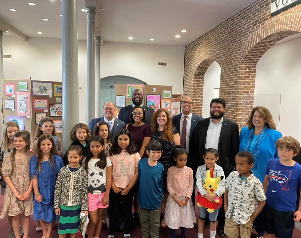 Mayor Wilson, members of City Council, and Alexandria students stand in front of a gallery of Earth Day-themed student art at the "We Are Eco-City" event.