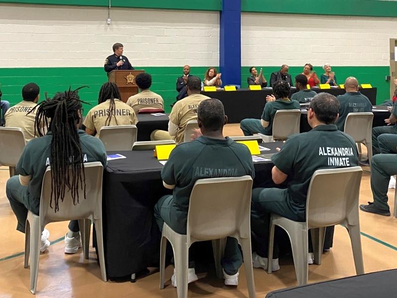 sheriff speaking at podium with several people seated and applauding inmates who are seated and wearing tan and green jumpsuits