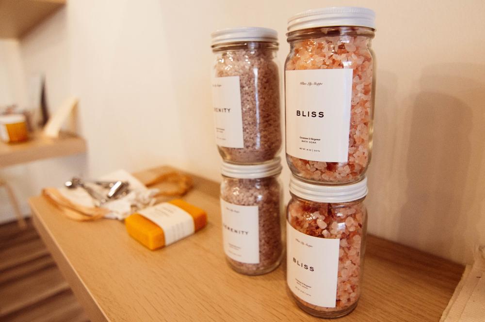 Four jars of bath soak are stacked two by two, all of them full of what looks like pink rock salt. Two of the jars are labeled Bliss and two are labeled Serenity. On the shelf beside the bath soak are a bar of soap and an out-of-focus silver utensil.