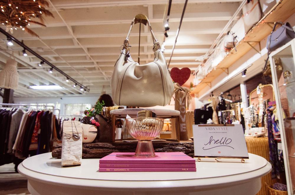 A round white table is decorated with stacked pink Alexandria shopping guides topped with a transparent pink glass dish. To the right of the stack is a small framed sign that says "hello". To the left is a piece of driftwood used to display a silver necklace. Behind the stack of guides is a pedestal displaying a white leather purse. In the background are racks of colorful clothes and a gold-framed mirror displaying jewelry. 