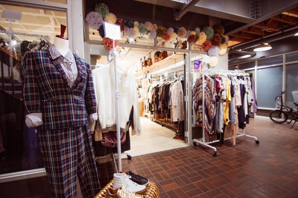 White clothing racks filled with colorful cloths frame the store's entrance. To the far left, closest to the camera, a mannequin is wearing a suit of blue, cream and teal plaid over a white blouse with blue polka dots. Above the store's entrance is a colorful banner of ruffled paper flowers