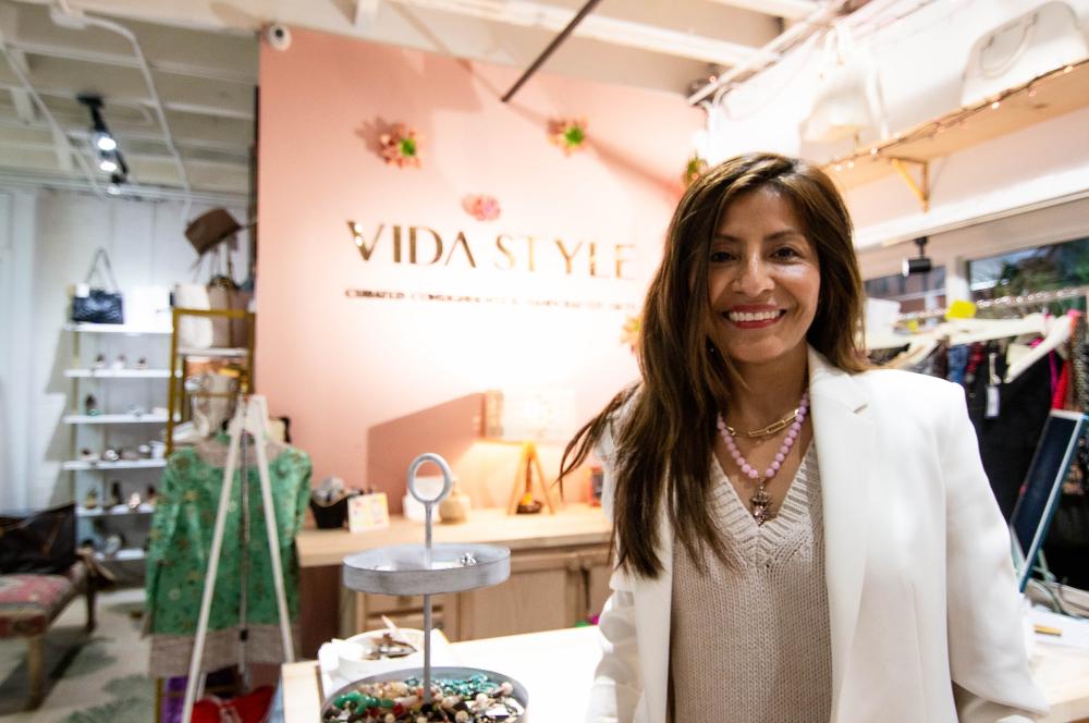 A brown haired woman poses for the camera in a cream-colored sweater and white blazer. Behind her are display racks of clothing and a pink wall with "Vida Style" in gold letters