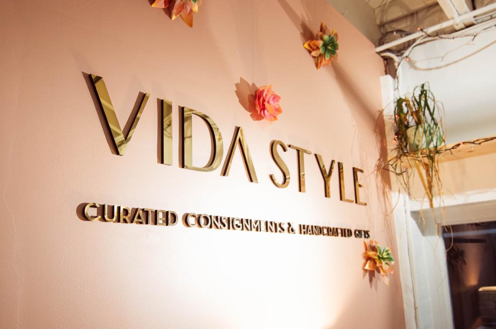 Gold lettering on a pink wall spells out "VIDA STYLE" and below that "Curated Consignment & Handcrafted Gifts". The wall is dotted with three-dimensional flowers in complementary colors.