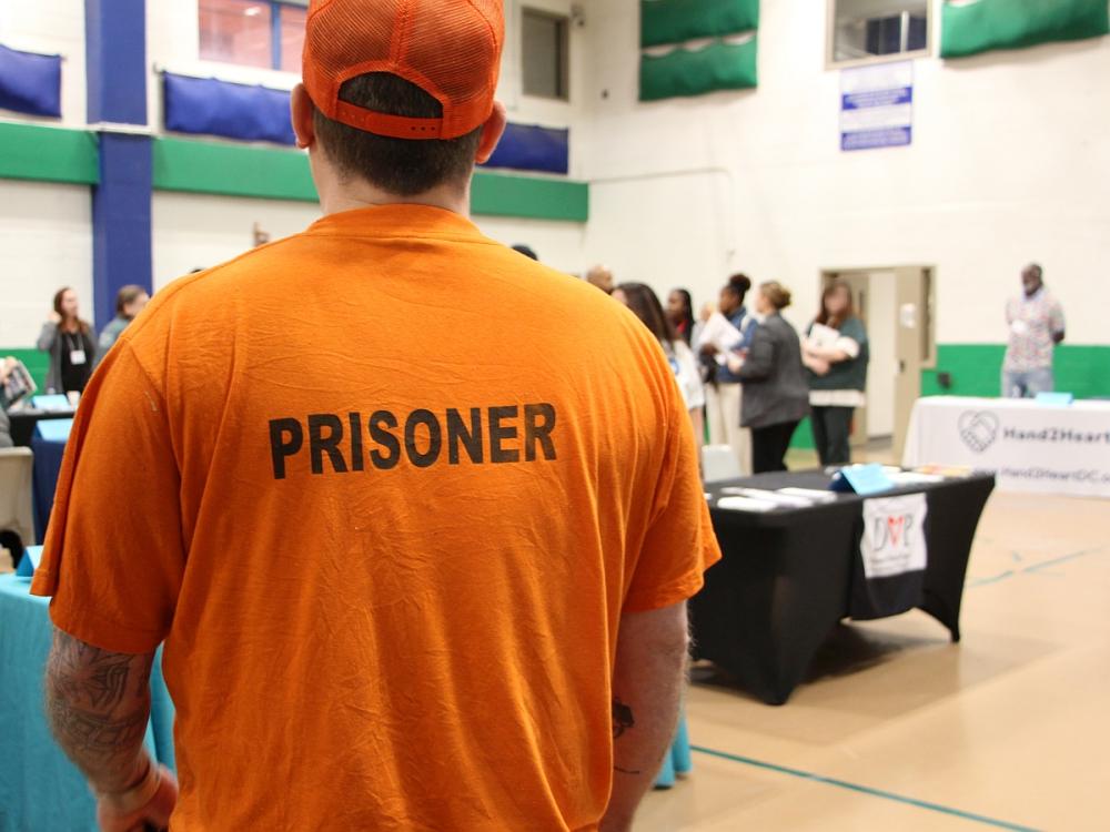 View from behind an inmate in an orange baseball cap and orange shirt with the world 'prisoner' on the back, as the inmate looks toward tables and people at a resource fair