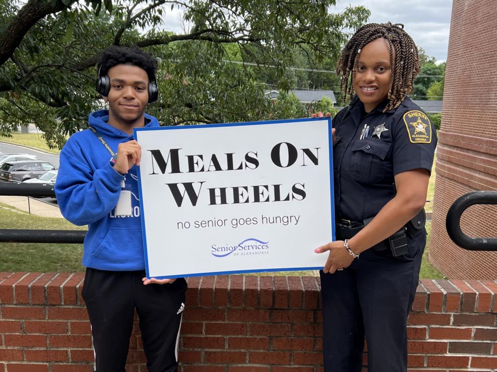 teen worker and deputy in uniform holding a sign that says Meals on Wheels