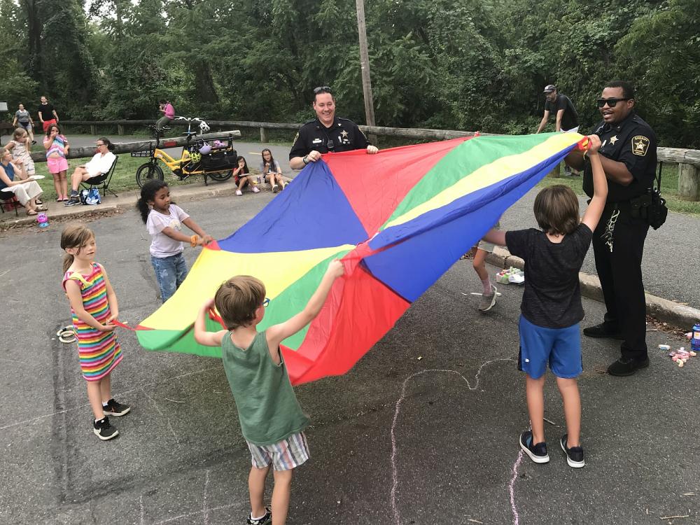 Two deputies and five children playing with a rainbow colored parachute on a playground during National Night Out