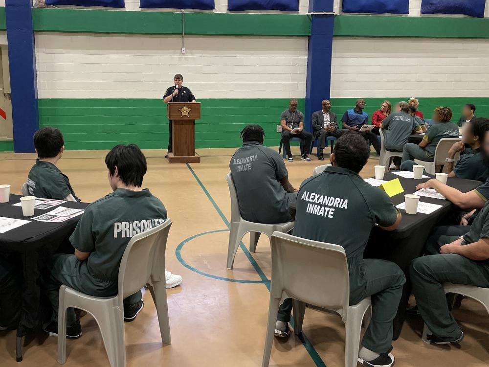 sheriff speaking at a podium to several inmates wearing green jumpsuits and seated at tables