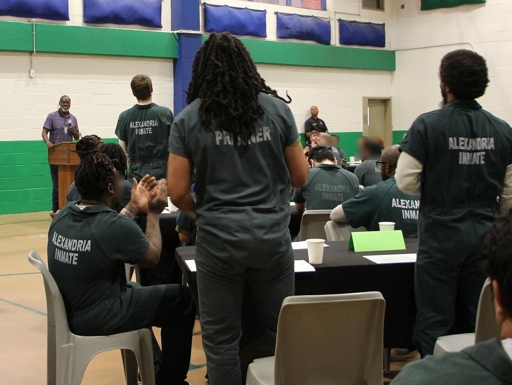 Several inmates wearing green jumpsuites and seen from the back, with some seated and three standing, at a recognition program.