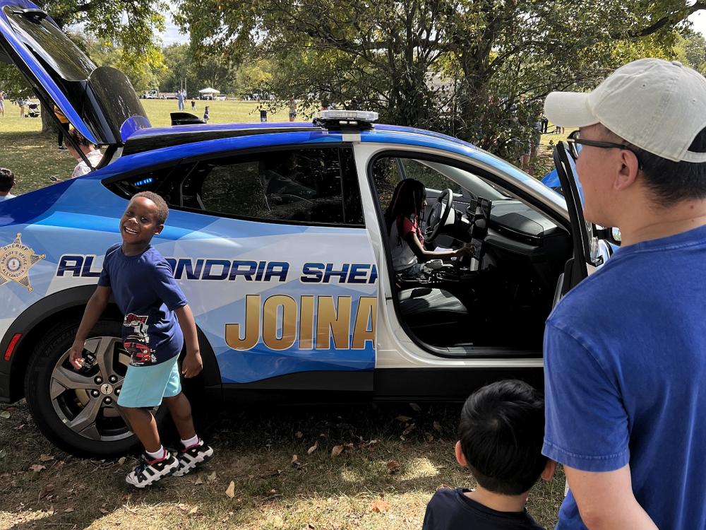 Child smiling outside blue sheriff's cruiser with adult and child looking on