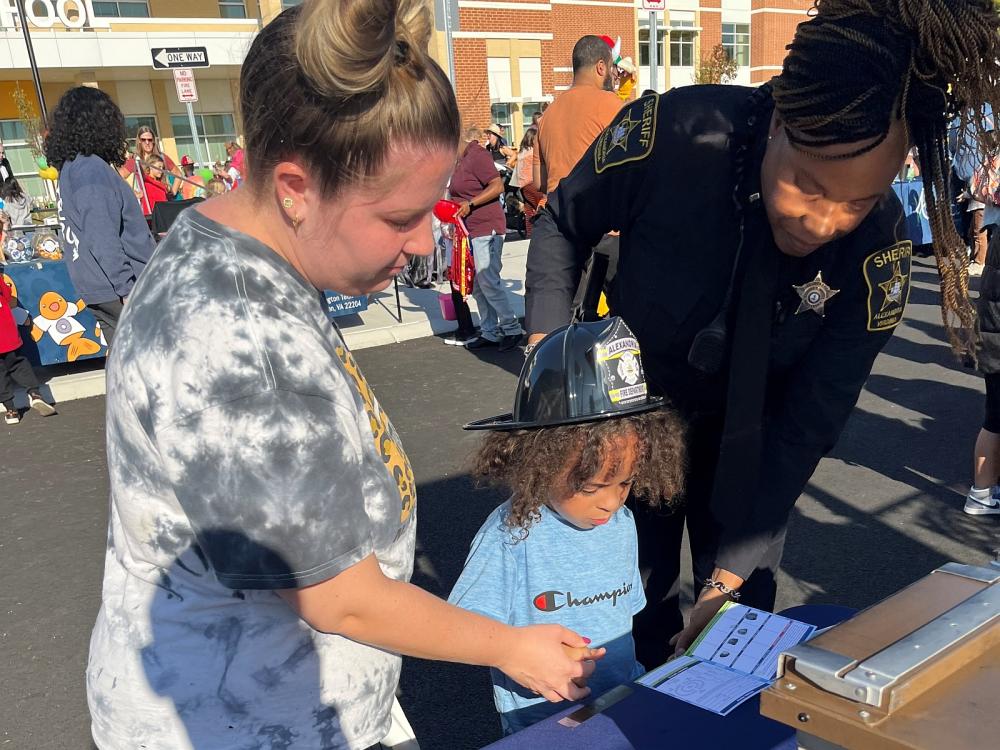 Civilian and deputy in blue uniform assisting a small child in getting fingerprinted at a community event.