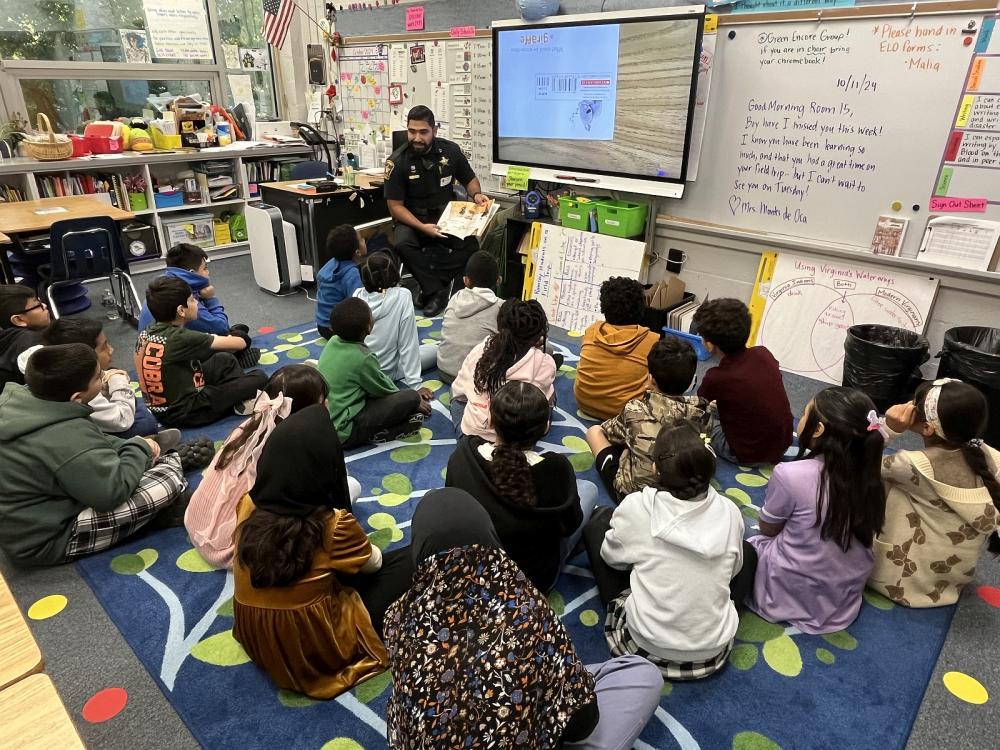 Deputy in blue uniform seated and reading a book to school children sitting on the floor of a classroom.