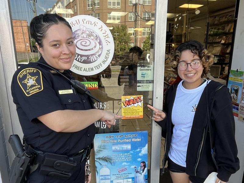 Deputy in blue uniform with teen next to her, both standing at doorway of a store and pointing to a yellow and orange window cling that says "Wait"