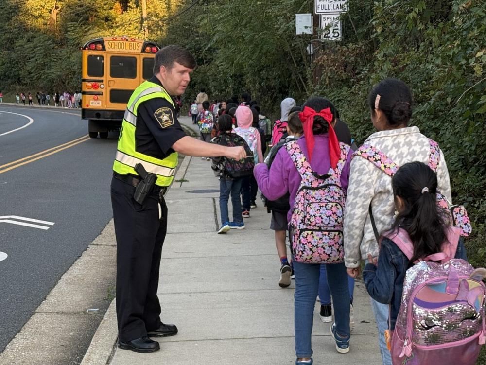 Several school children walking down a sidewalk away from the camera and Sheriff in blue uniform and yellow safety vest exchanging fist bump with one of the kids. A school bus is visible in th background.