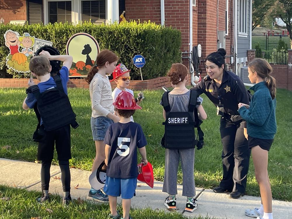 Deputy talking with several children outside a home with two kids trying on black vests, including one that says "Sheriff"