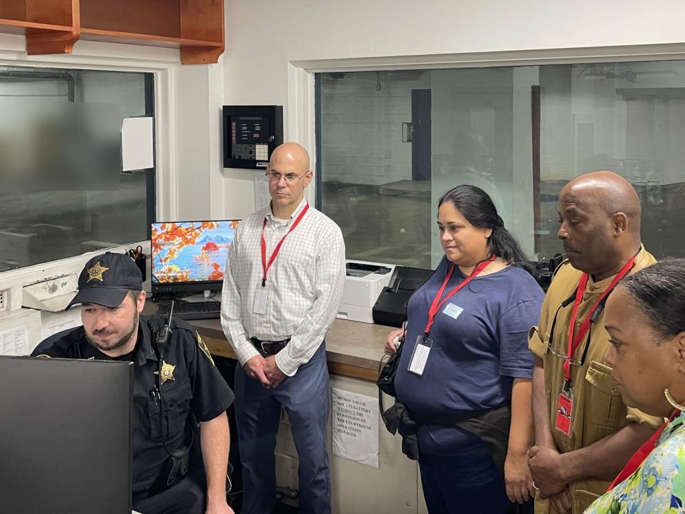 Deputy in blue uniform seated and looking at computer screens while four civilians look on and listen