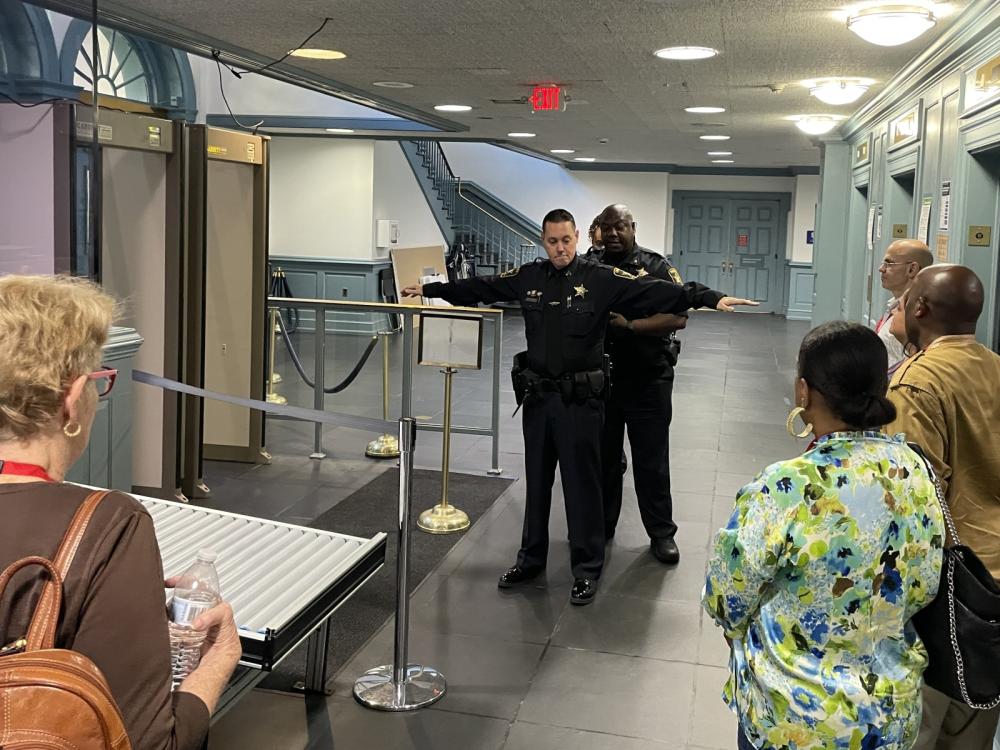 Inside a couthouse, two deputies in blue uniforms demonstrating how to scan and search someone after passing through a magnatometer while five citizens look on