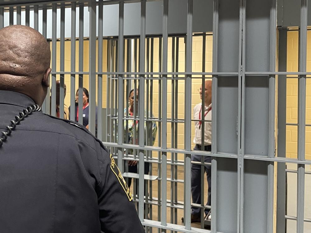 Four individuals with metal cell bars in front of them and a deputy, partially seen from behind, looking at them