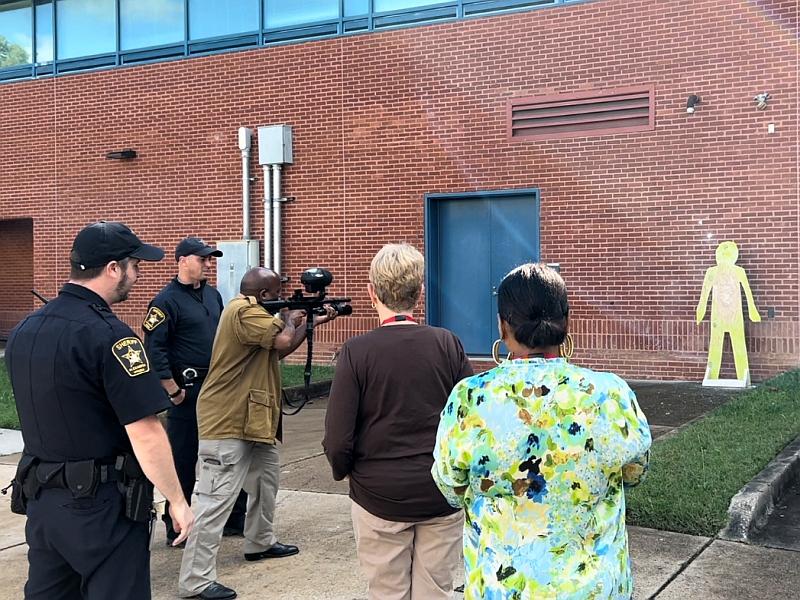 Deputy in blue uniform observing citizen shooting a pepperball training round at a yellow cut out against a brick wall while another deputy and two other citizens look on