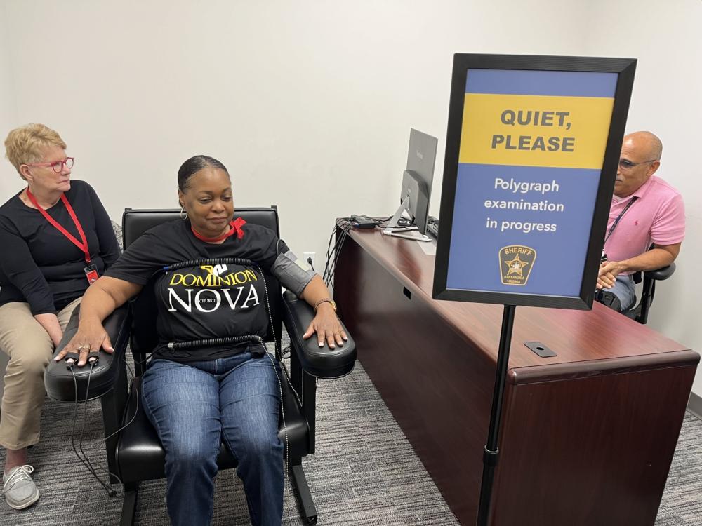 Two women sitting in chairs, one of them with cords attached to her hands and body, with an investigator seated at a desk looking at a computer screen, and a sign reading Quiet please, polygraph examination in progress