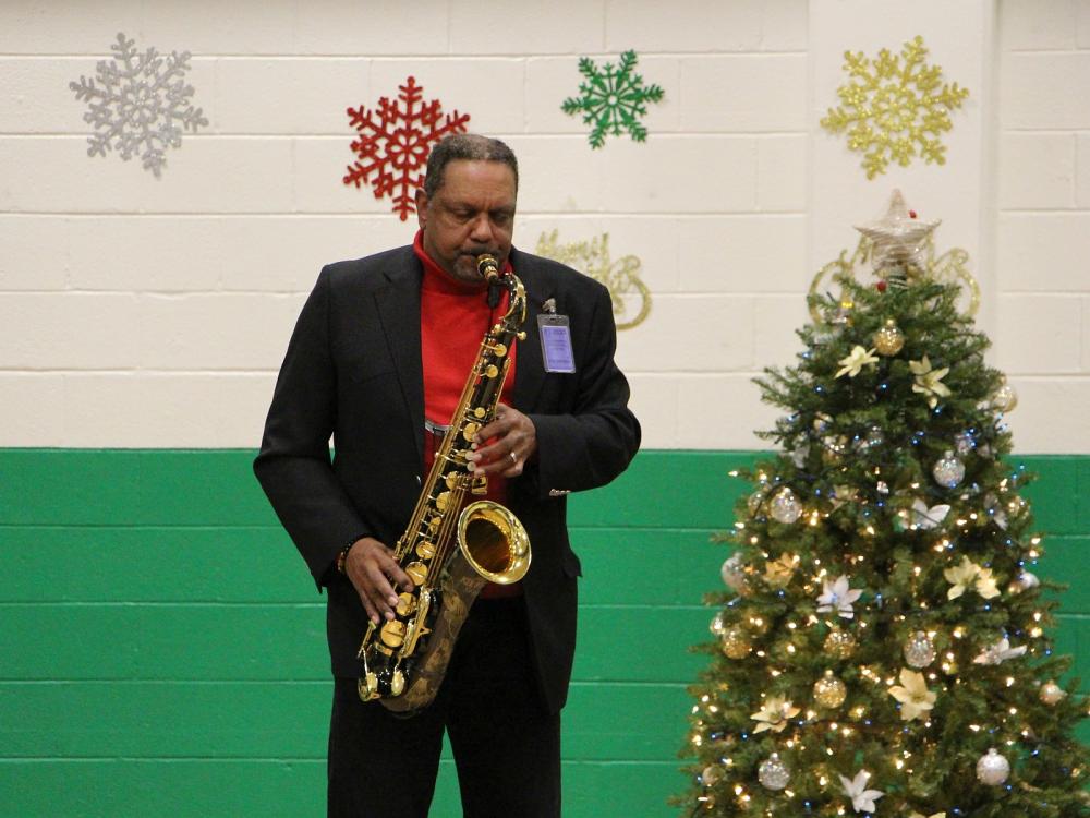 man wearing a black suit and red shirt playing the saxophone with a Christmas tree and holiday decorations in the background