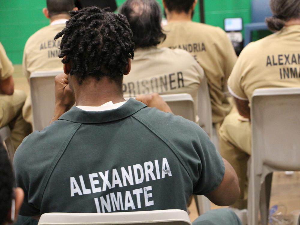 Inmate in a green jumpsuit, seated and only visible from behind, holding his hands up as he listens to music, with other inmates wearing tan jumpsuits seated nearby