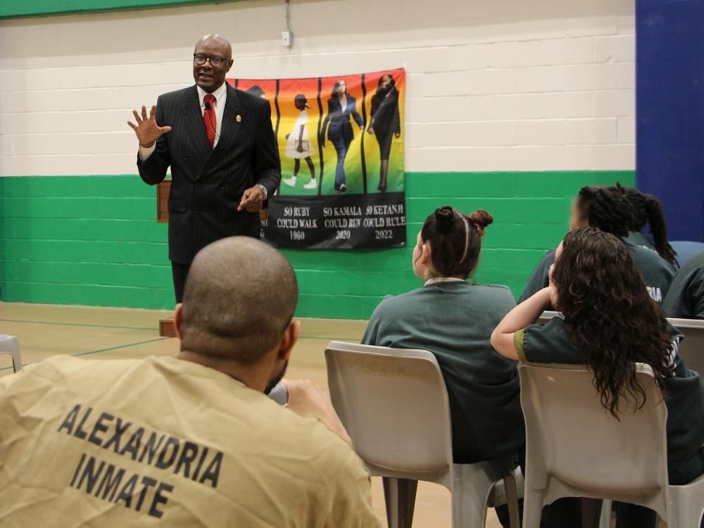 guest speaker wearing a business suit answer a question from an inmate wearing a tan jumpsuit that reads "Alexandria Inmate" on the back