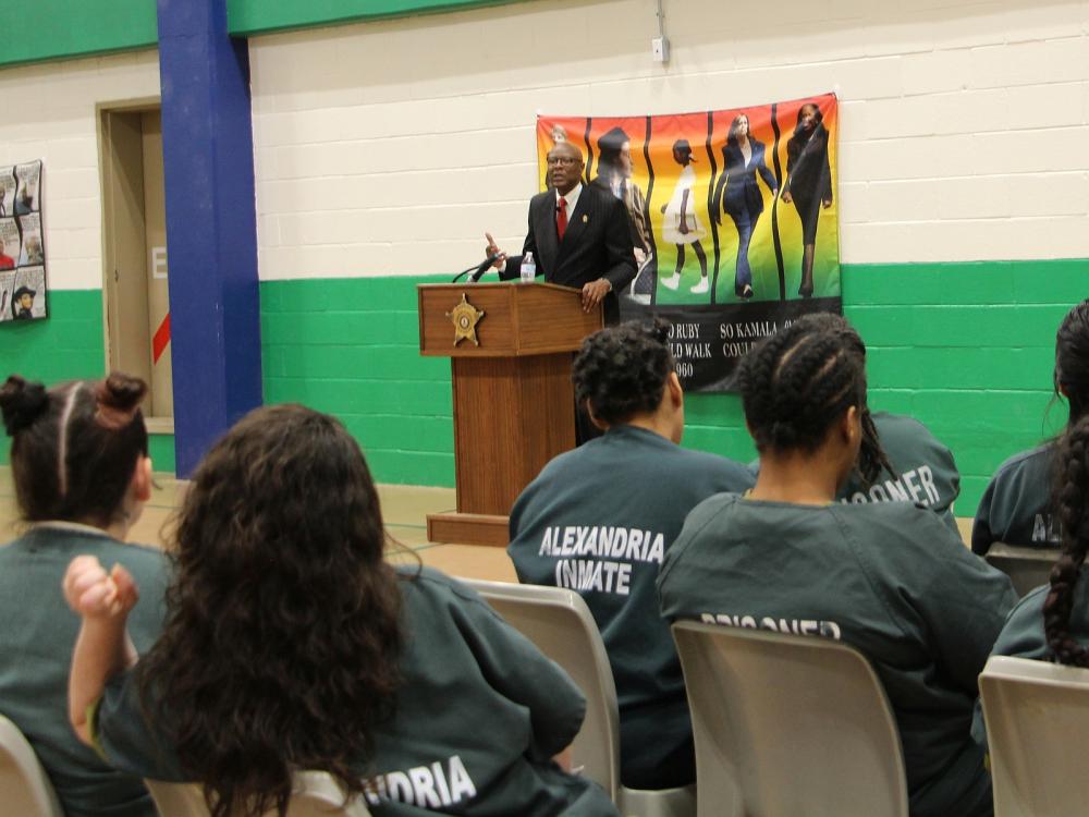 Man in a suit standing at a podium and speaking to several inmates who are seated at tables, wearing green jumpsuits and only visible from behind