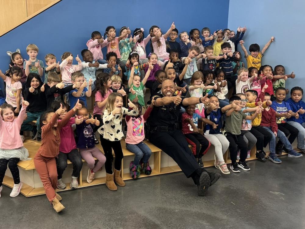 Deputy in a blue uniform with about 30 small children around him inside a school, all giving thumbs up