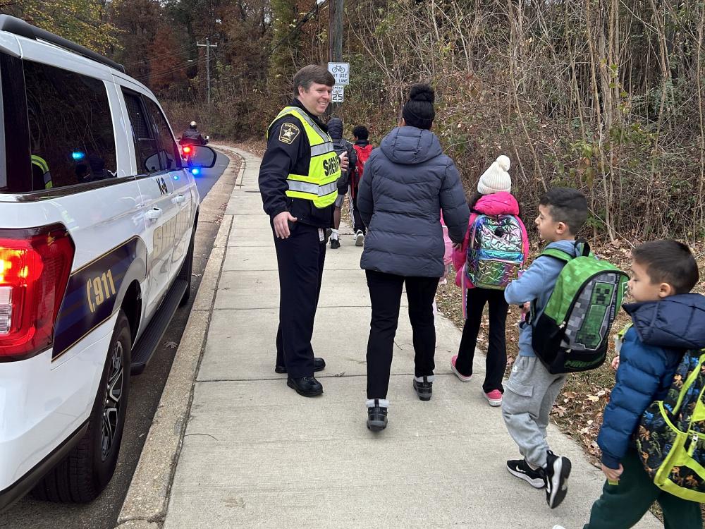 Sheriff in blue uniform and bright yellow traffic vest watching as several small children and an adult walk to school down a sidewalk. A white law enforcement SUV with its lights on is parked to the left.
