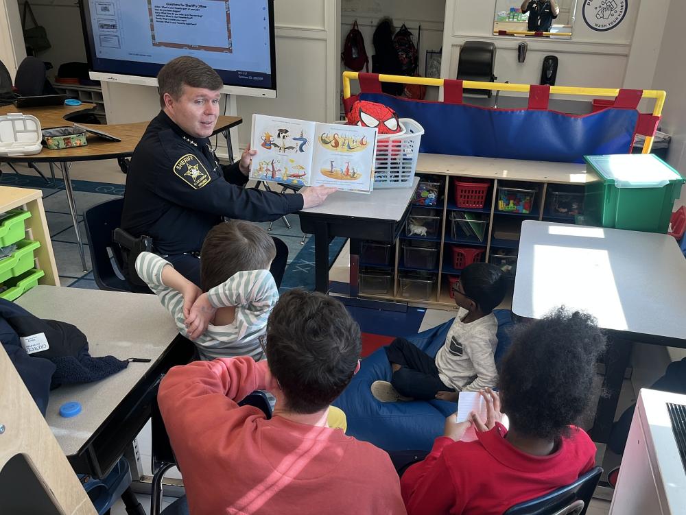 Sheriff in a blue uniform seated and reading an illustrated book to small children in a classroom